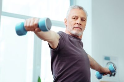 Strong fit aged man looking at his hand while training with dumbbells