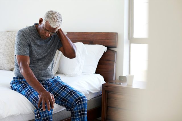 Senior man suffering from neck pain sitting by his bed at home