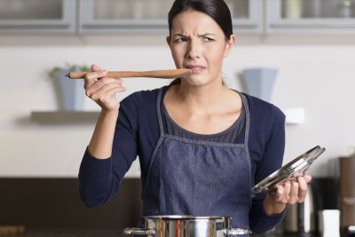 female cook standing at the hob in her apron tasting her food in the saucepan with a grimace as she finds it distasteful and unpalatable