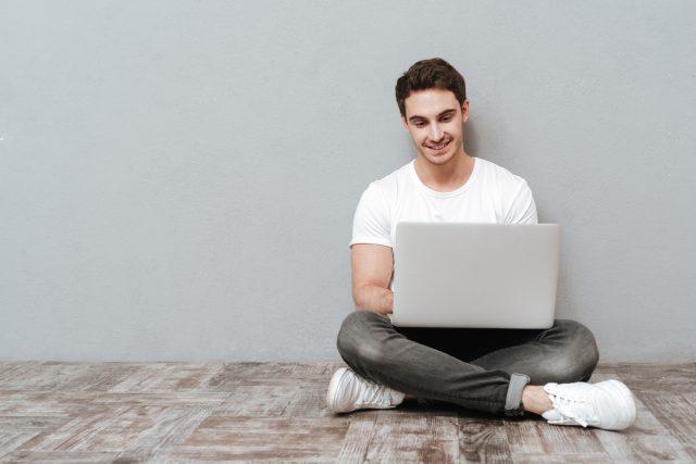 Man sitting on the floor with laptop computer in studio. Isolated gray background