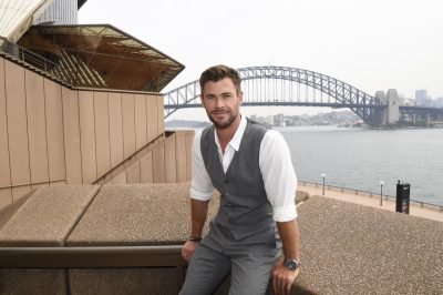 chris hemsworth in gray vest, gray pants, and white shirt sitting on a ledge at sydney opera house