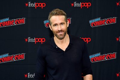 ryan reynolds in black unbuttoned henley in front of ny comic-con step and repeat