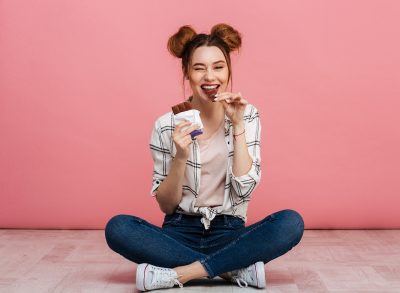 woman sitting in front of pink wall eating chocolate bar