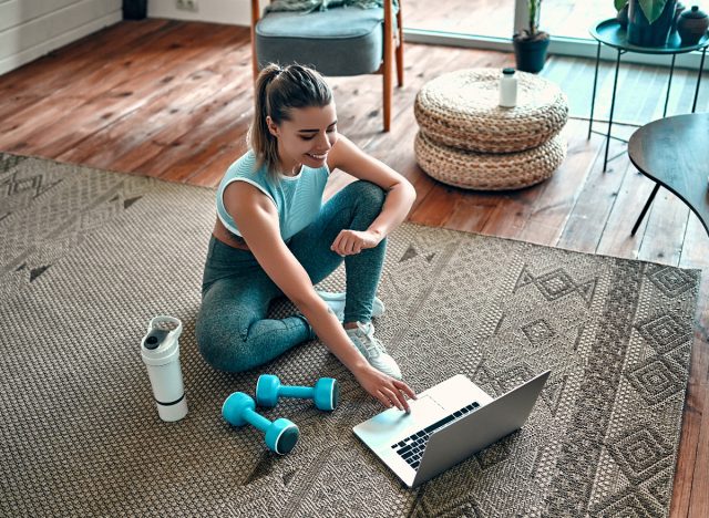 woman sitting on floor with dumbbells, protein shake, and laptop