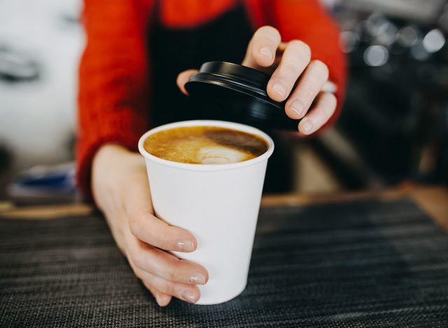 Barista in apron is holding in hands hot cappuccino in white takeaway paper cup. Coffee take away at cafe shop