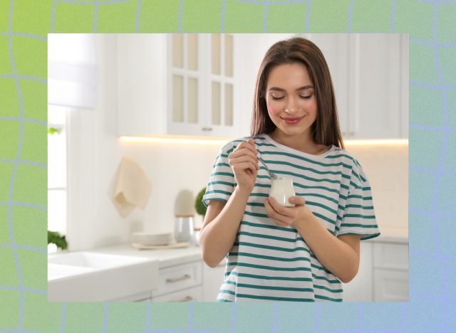 happy woman in green and white striped shirt eating greek yogurt out of small jar in bright white kitchen