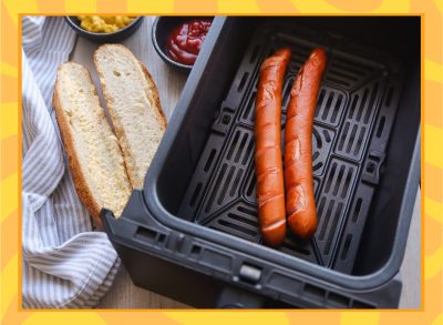 two hot dogs sitting in air fryer basket with bun and condiments on the table next to it