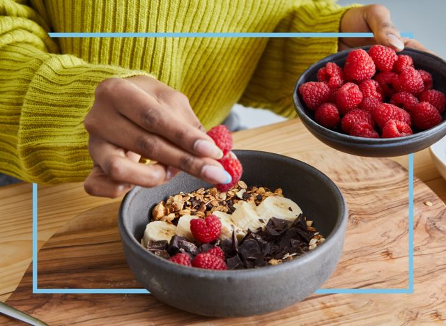 woman putting raspberries on top of a bowl of yogurt with chocolate chips and bananas