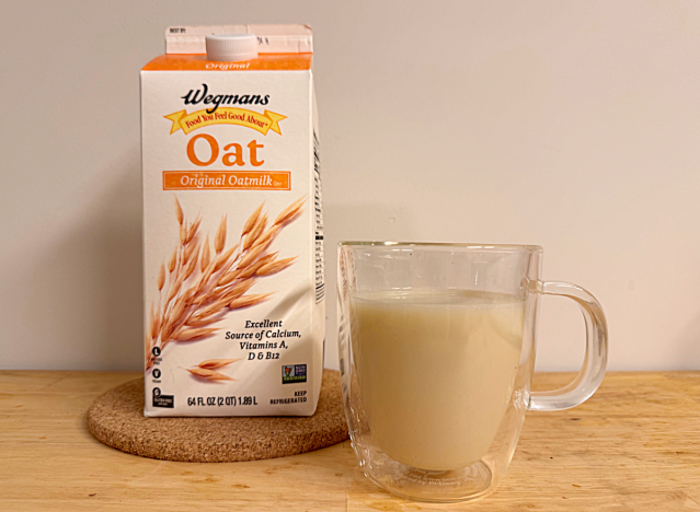 a container of wegman's oat milk next to a glass mug filled with coffee and oat milk.