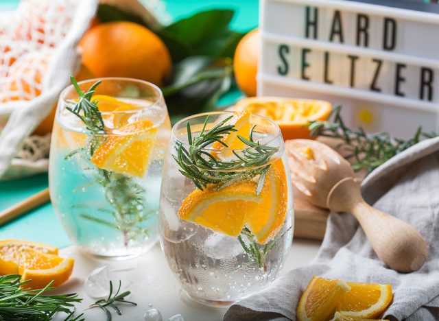 Hard seltzer cocktail with orange, rosemary and ice on a table. Summer refreshing beverage, drink with trendy zero waste accessories, bamboo straw and mesh bag.