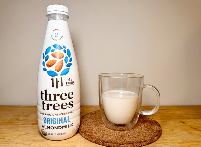 a container of three trees almond milk next to a glass of it 