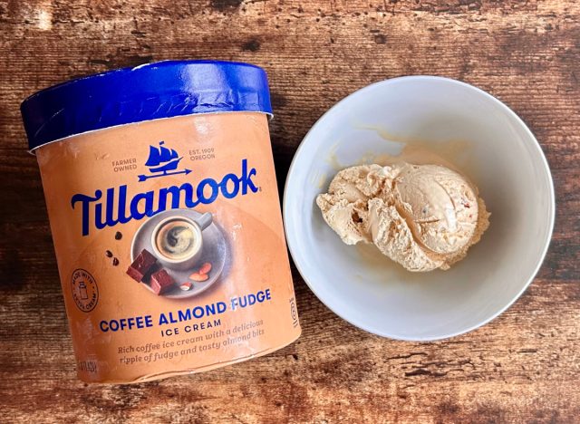 a container of tillamook's coffee almond fudge ice cream next to a bowl of breyers coffee ice cream on a wooden table background