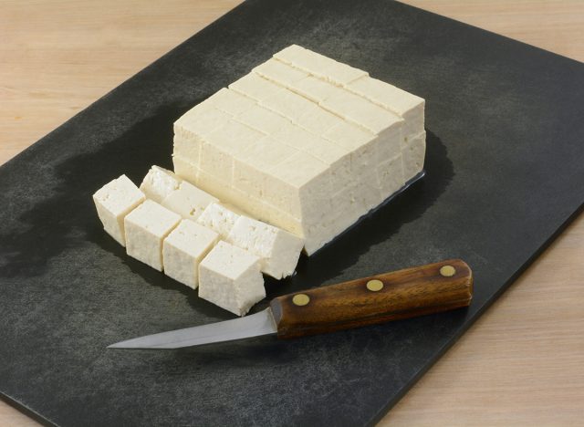 Block of extra firm tofu cut into cubes on cutting board with kitchen knife