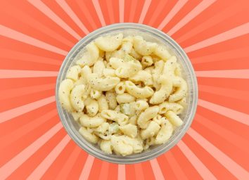 A round open container of macaroni salad set against a vibrant reddish background.