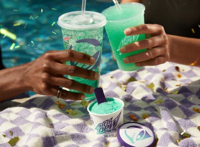 Hands holding Taco Bell Baja Blast and Baja Blast Freeze, with a cup of Baja Blast Gelato sitting nearby on a table