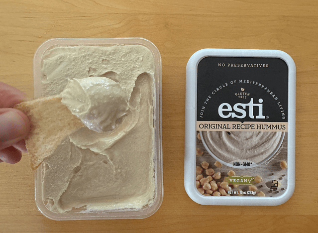 esti hummus container open with someone dipping a pita chip into it 