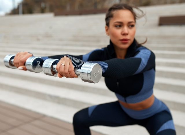 Sporty girl jumping with dumbbells. Photo of a sportswoman with a perfect body against a city background. Strength and motivation. squat with a load