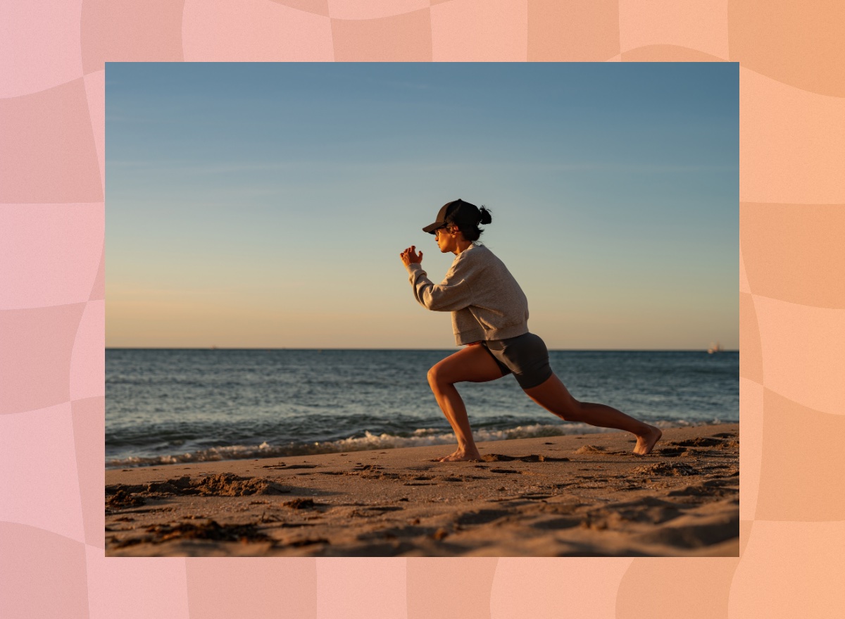 fit woman in biker shorts, a sweatshirt, and a baseball cap doing walking lunges on the beach at sunset