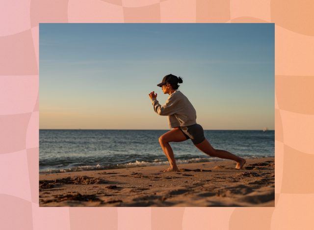 fit woman in biker shorts, a sweatshirt, and a baseball cap doing walking lunges on the beach at sunset