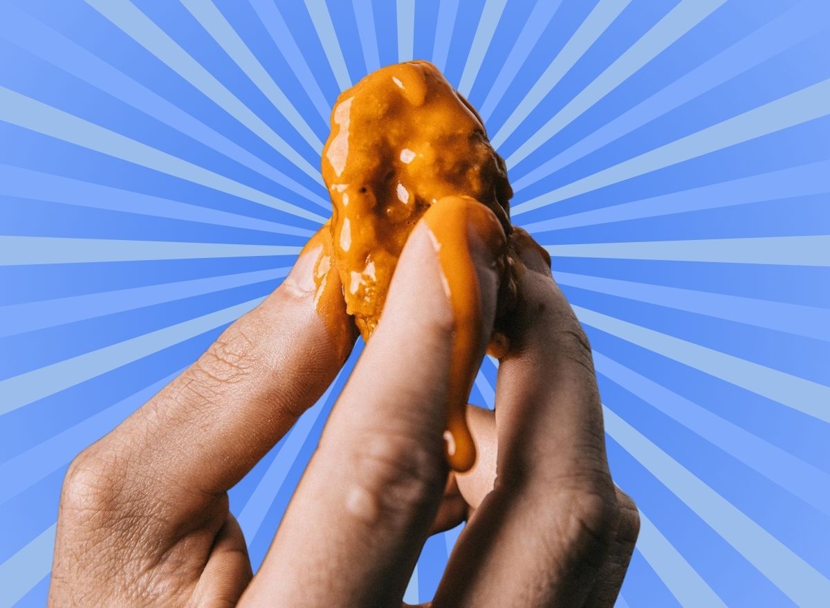 A saucy boneless chicken wing held aloft by the fingertips, set against a vibrant blue background.