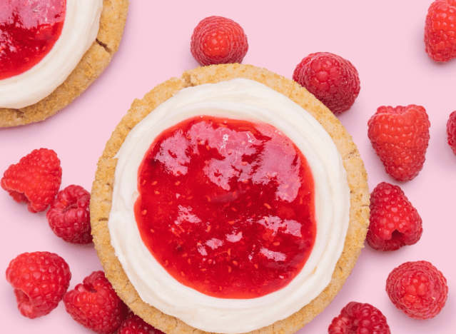 crumbl raspberry cheesecake cookie with raspberries around it on a pink background