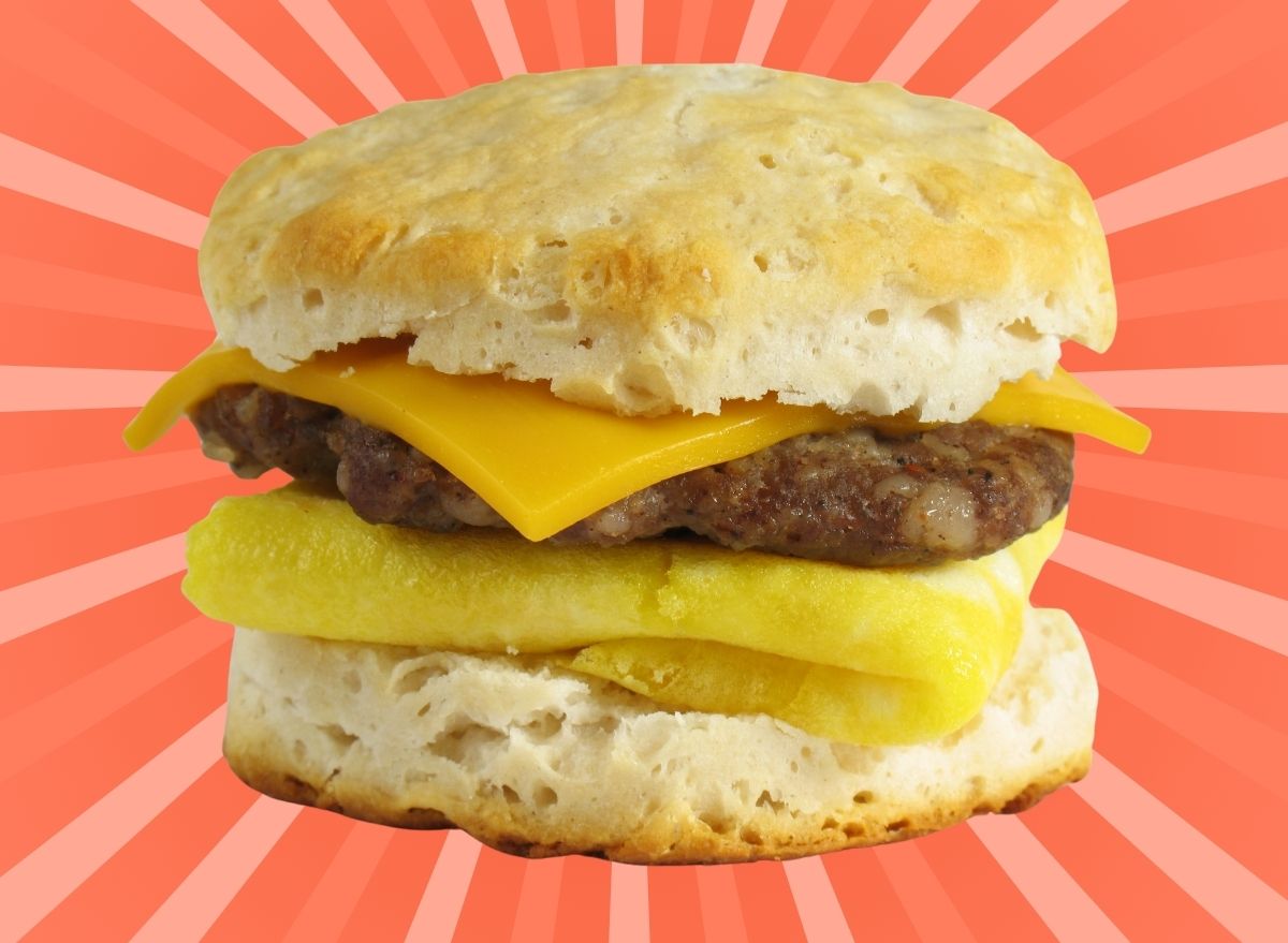 A fast food breakfast sandwich with egg, sausage and cheese on a biscuit, set against a vibrant orange background