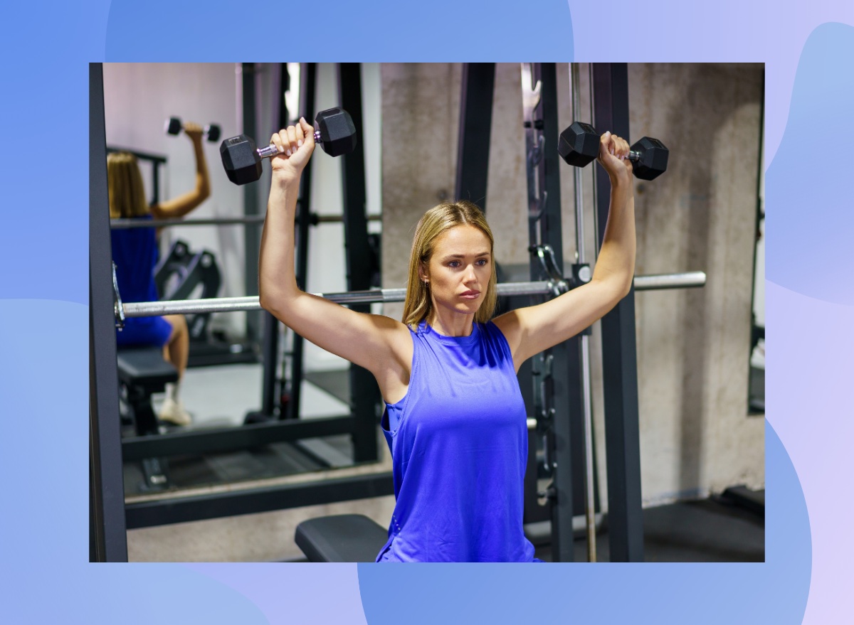 focused blonde woman doing seated overhead press with dumbbells in the weight room at the gym