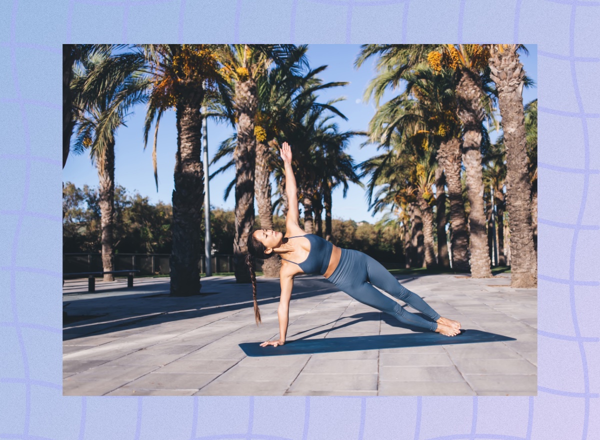 focused woman wearing blue sports bra and leggings doing side plank on yoga mat surrounded by palm trees