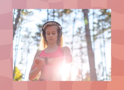 blonde woman wearing headphones and pink sports bra checking fitness tracker while on a run through the woods on sunny day