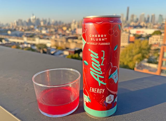 A can of Alani Cherry Slush beside a glass of the energy drink, perched on a rooftop overlooking the NYC skyline.