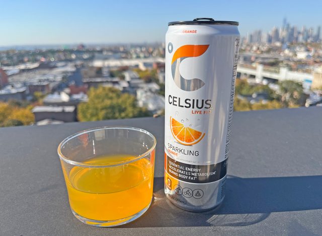 A can of Celsius Sparkling Orange energy drink beside a glass of the beverage, perched on a rooftop overlooking the NYC skyline.