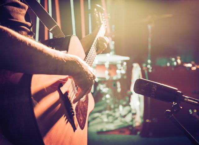 An acoustic guitar close-up in a concert hall, with a drum set in the background.