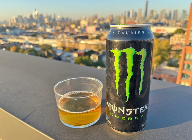 A can of the original Monster energy drink beside a glass of the beverage, perched on a rooftop overlooking the NYC skyline.