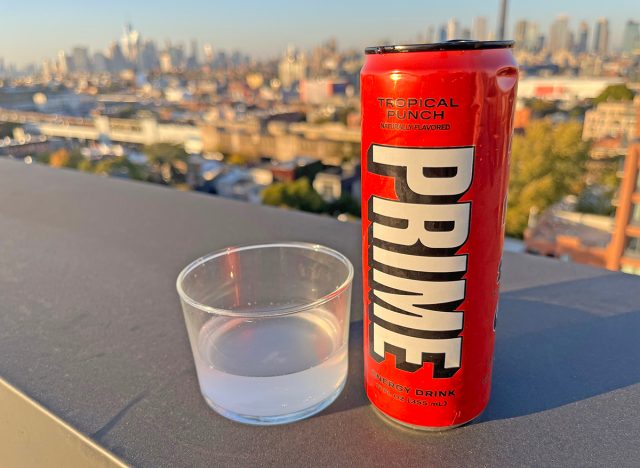 A can of Prime Tropical Punch beside a glass of the energy drink, perched on a rooftop overlooking the NYC skyline.