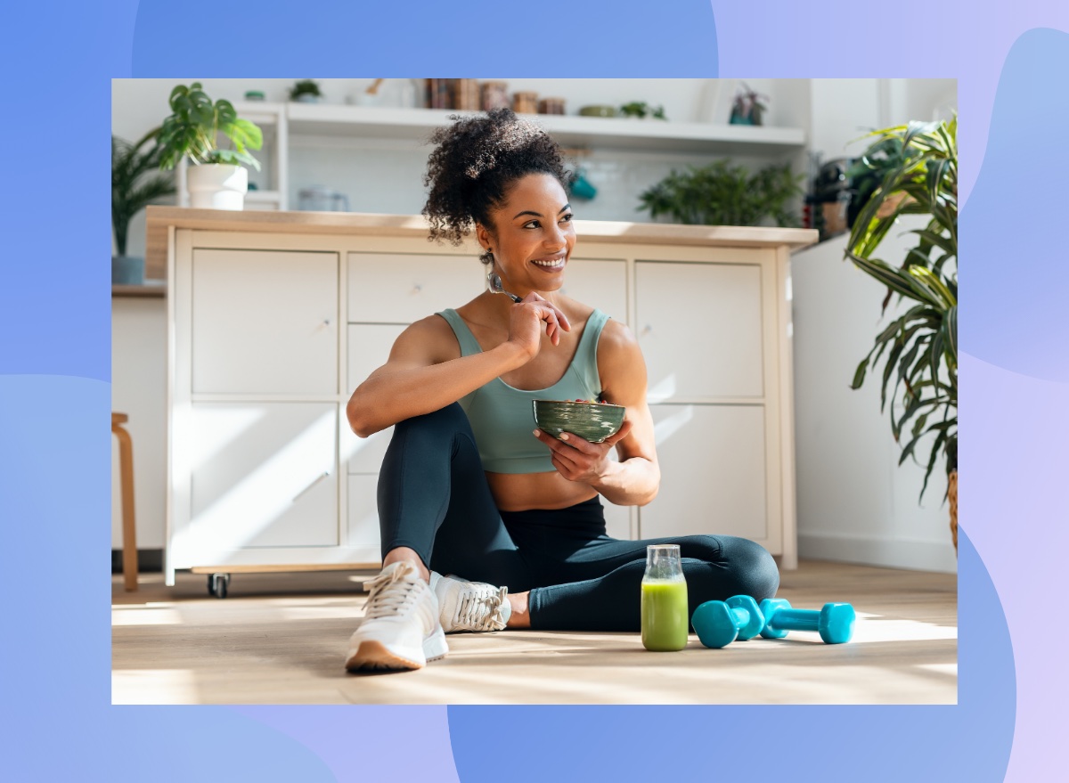 happy fitness woman sitting on floor eating healthy breakfast next to green juice and dumbbells in bright kitchen