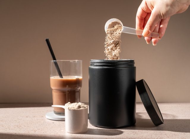 Woman holding scoop of protein powder above container with a protein shake in the background