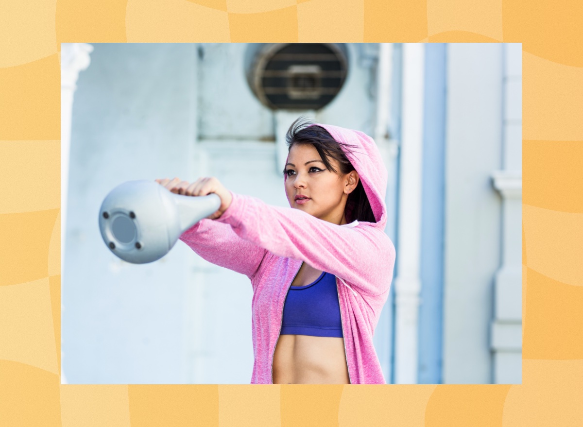 fit, determined brunette woman in pink hoodie and purple sports bra doing kettlebell swing exercises outdoors