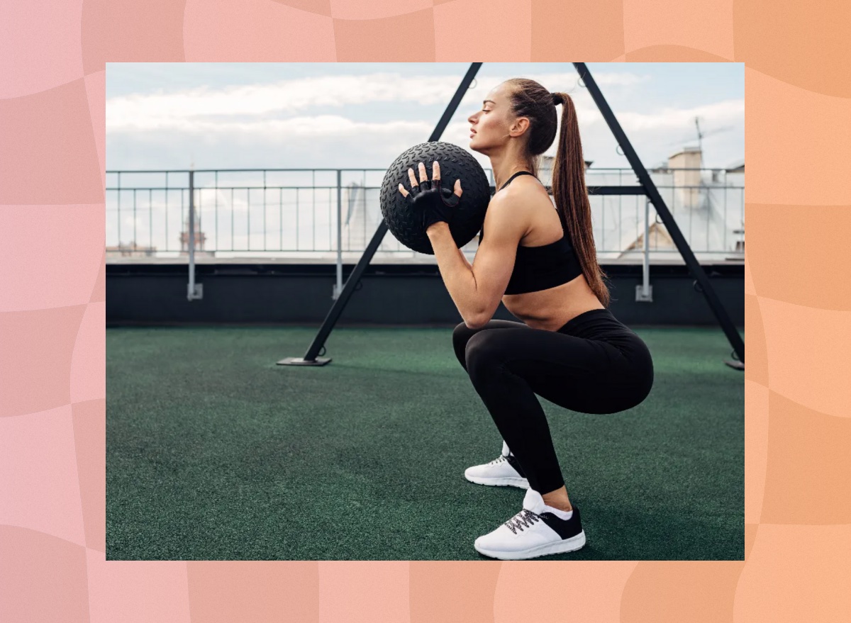 determined brunette fitness woman doing medicine ball squats on turf rooftop gym