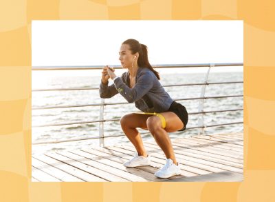 fit brunette woman doing resistance band squat exercise on boardwalk by the ocean