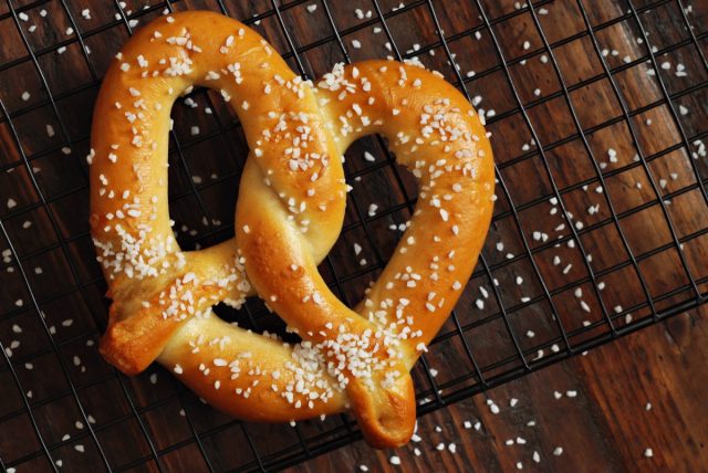 Freshly baked soft pretzel with generous sprinkling of coarse salt on wire cooling rack over rustic dark wood.