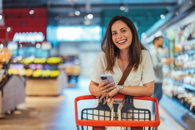 Portrait of happy woman buying groceries in supermarket and looking at camera. Purchasing Goods with Smartphone at Grocery Store. Female customer shopping with smartphone checklist