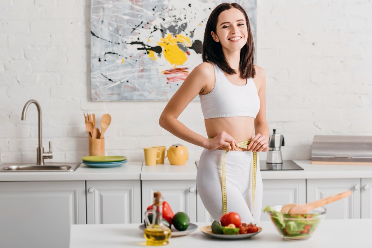 Selective focus of fit woman smiling at camera while measuring waist with tape near salad and fresh vegetables on table.