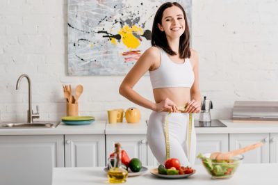 Selective focus of fit woman smiling at camera while measuring waist with tape near salad and fresh vegetables on table.