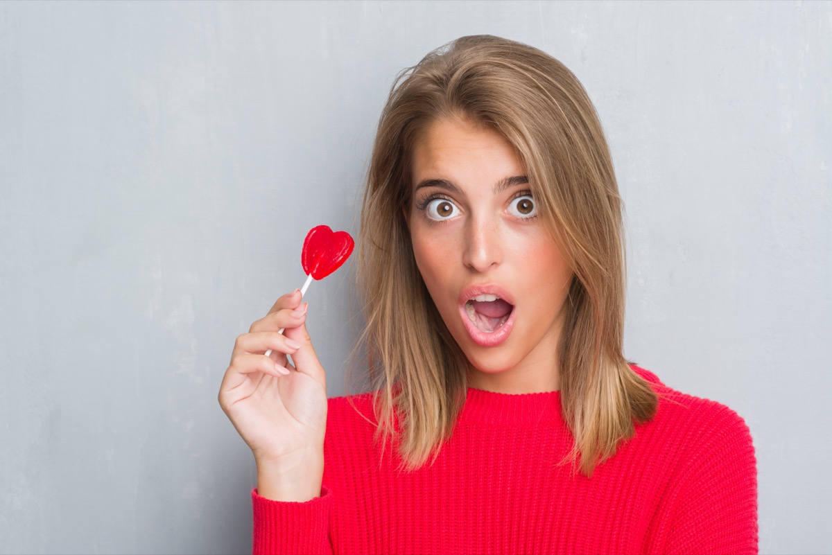 Beautiful young woman over grunge grey wall eating red heart lollipop candy scared in shock with a surprise face, afraid and excited with fear expression