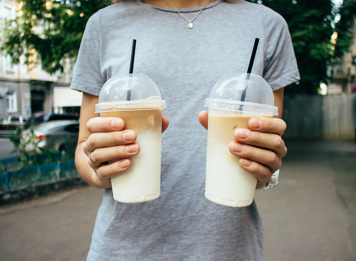 Girl holding two coffee lattes in plastic transparent cups with tubes. Female offering cold caffeine refreshments standing at city street at summer day.