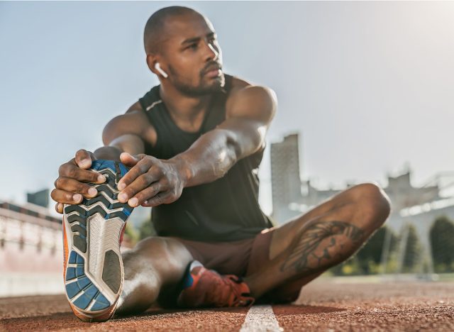 Sporty muscular african male athlete in earphones stretching legs while sitting at the stadium race track