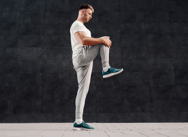 Full length of attractive Caucasian bearded blonde man standing on one foot and stretching leg. In background is gray wall.