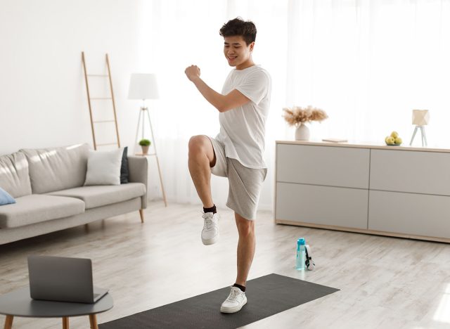 Online Workout Concept. Cheerful Smiling Asian Guy Training In Living Room, Doing Twist Knees To Elbow Exercise. Happy Man Standing And Lifting Leg Up, Using Laptop Watching Tutorial Lookign At Screen