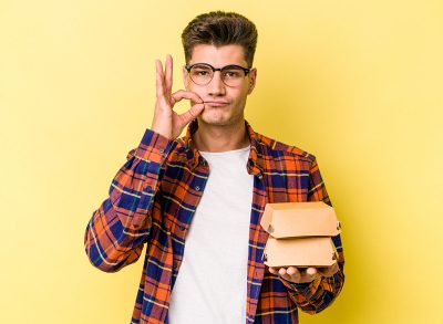 Young caucasian man holding a burger isolated on yellow background with fingers on lips keeping a secret.