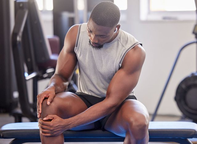 Black man, fitness and knee injury in gym after workout, training or intense exercise. Earphones, health and bodybuilder male from Nigeria suffering from sore leg, muscle pain or joint inflammation.